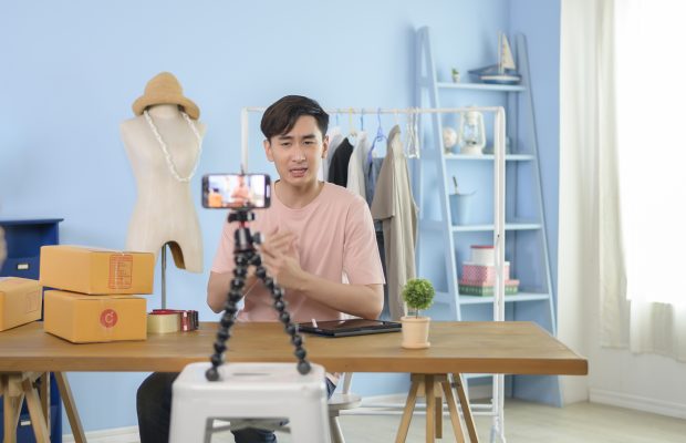 Un homme présente des vêtements devant un smartphone lors d’un live streaming dans sa boutique. Concept de commerce en ligne et de technologie numérique.
